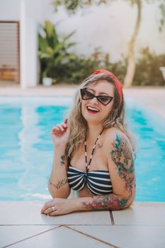 Tattooed woman in bikini laughing by a pool, enjoying a sunny day with sunglasses on.