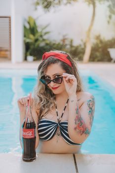 Young woman in sunglasses enjoying a refreshing Coca-Cola by the pool on a summer day.