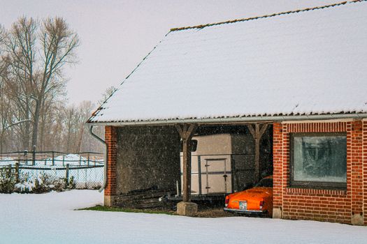 Vintage car parked in a snow-covered red brick garage during a winter snowfall.