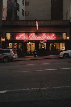 A bustling city street at night featuring a neon-lit restaurant front with parked cars.