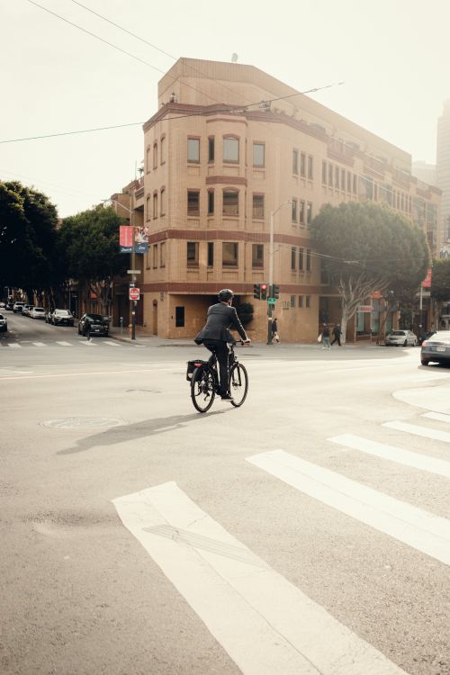 Free A man rides a bicycle across a sunlit city intersection, showcasing urban lifestyle and transport. Stock Photo