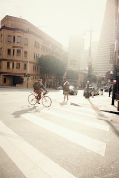A cyclist rides through an urban crosswalk as sunlight filters through city buildings.