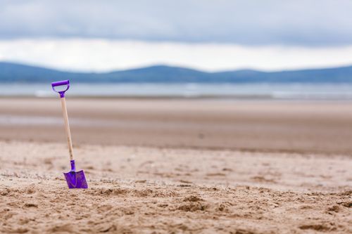 Free A purple shovel stuck in the sand on a beach with mountains in the background, under a cloudy sky. Stock Photo