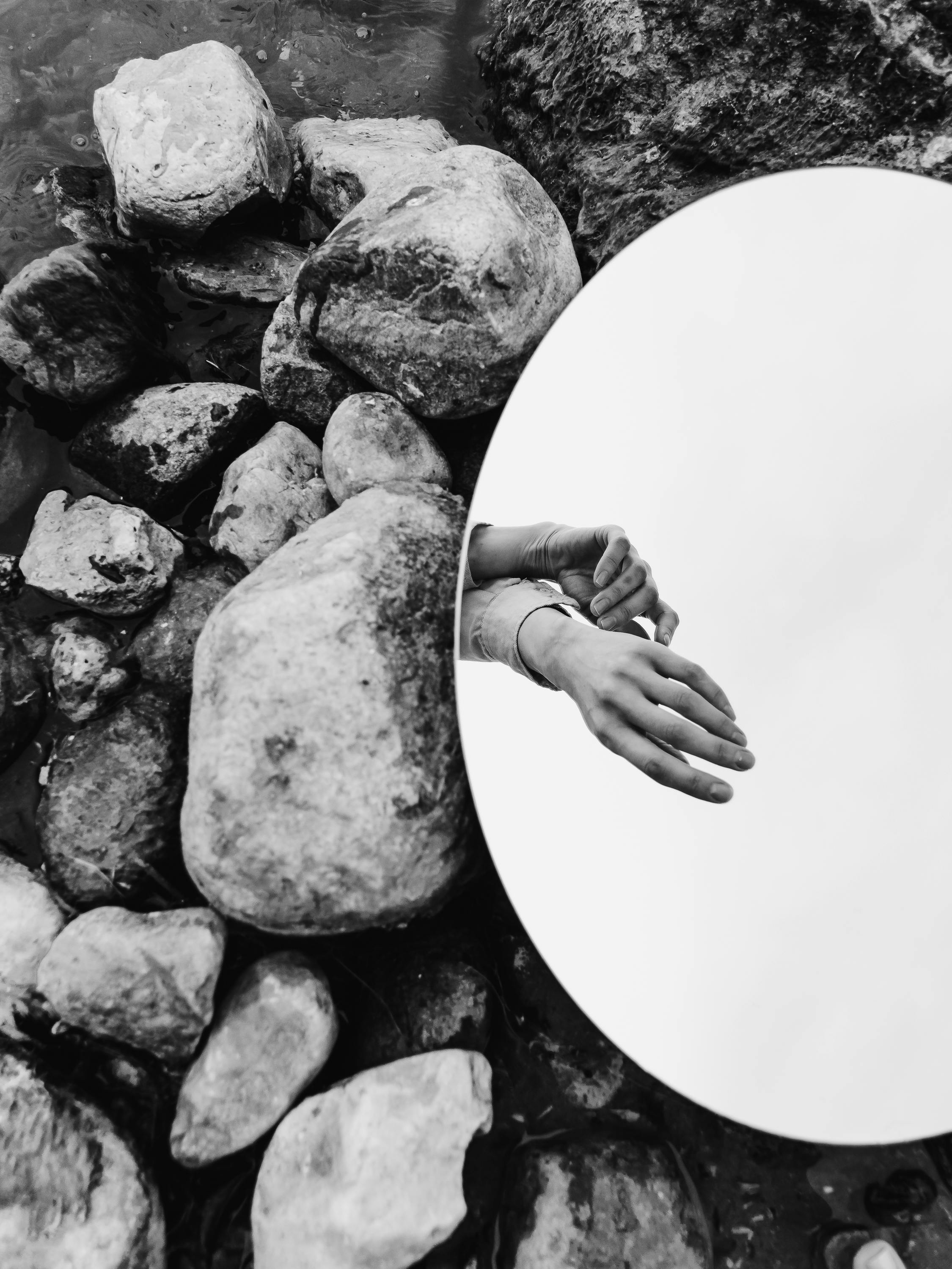 Black and white artistic shot of hands reflected in a mirror among river stones.