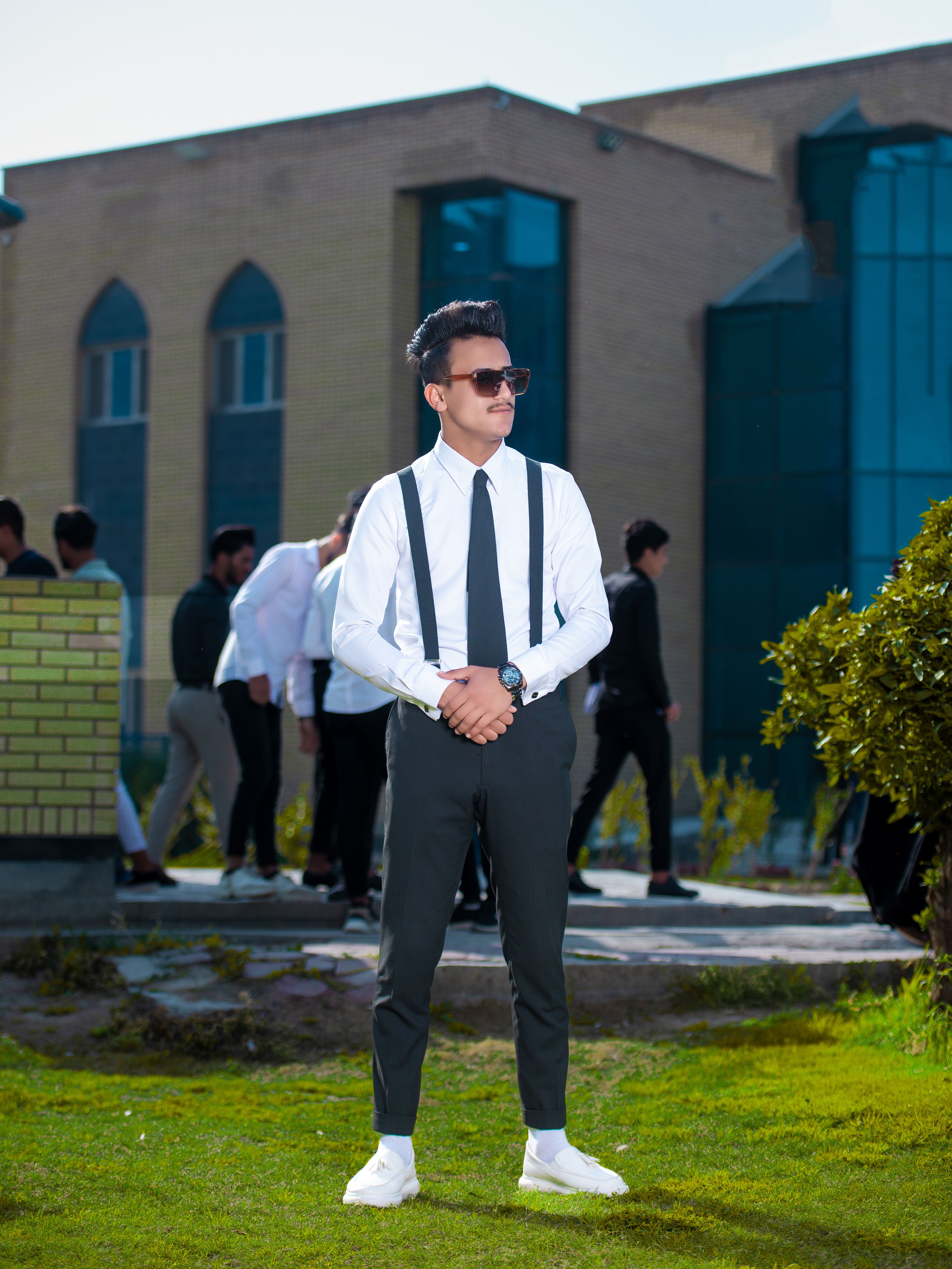 A fashionable young adult wearing suspenders and sunglasses poses confidently in an outdoor setting.