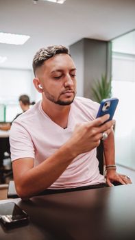 A young man with short hair uses his smartphone in a modern office setting, sitting at a desk.
