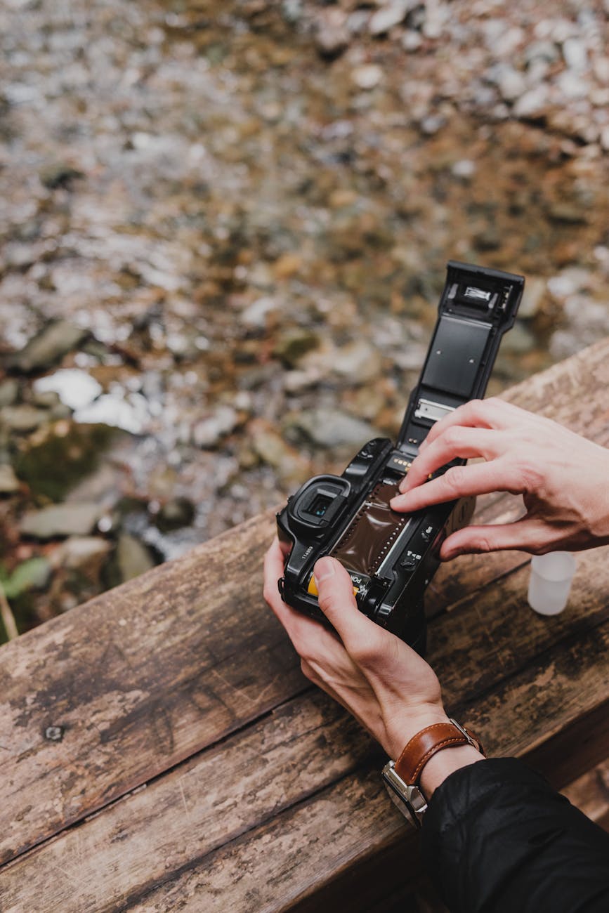 Close-up of hands loading film into an analog camera on a wooden railing by a stream.