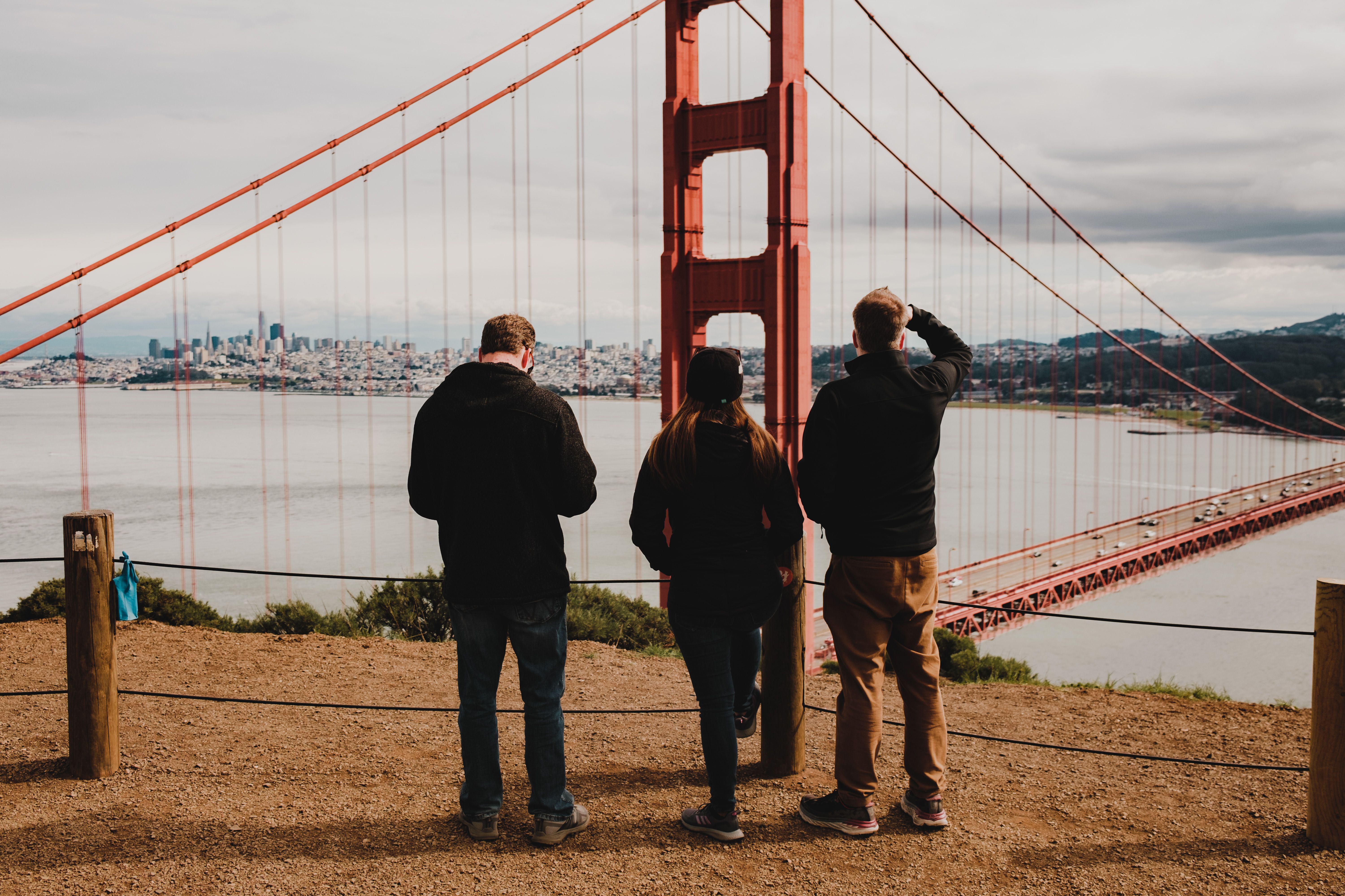 Three individuals admire the Golden Gate Bridge from a scenic viewpoint on a cloudy day.