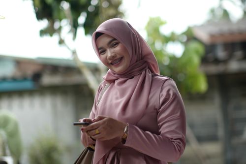 Free A cheerful woman in a pink hijab smiles while using her smartphone outdoors, capturing a moment of joy and connectivity. Stock Photo