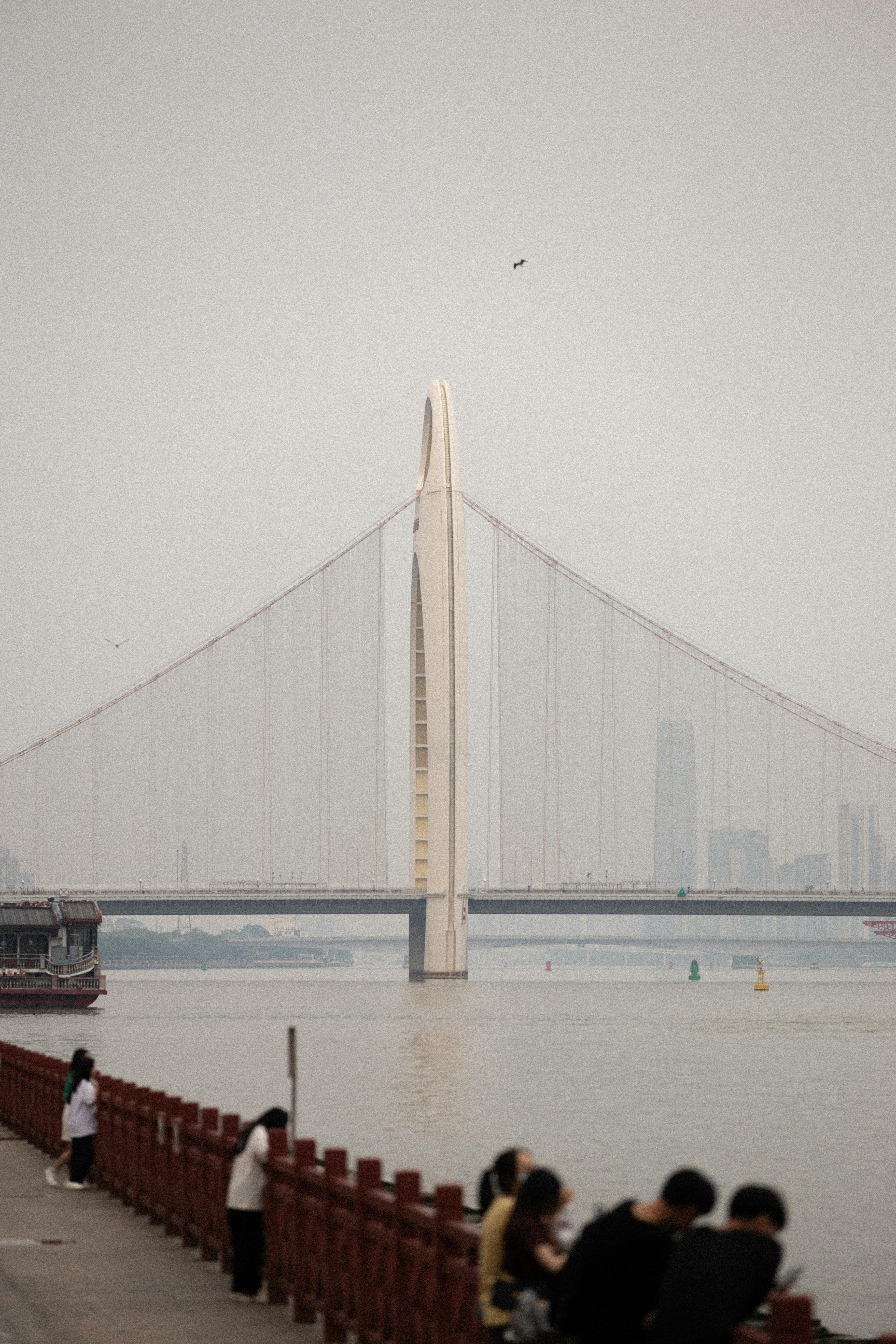 A modern cable-stayed bridge spanning a river with city skyline in the background, featuring people walking along a promenade