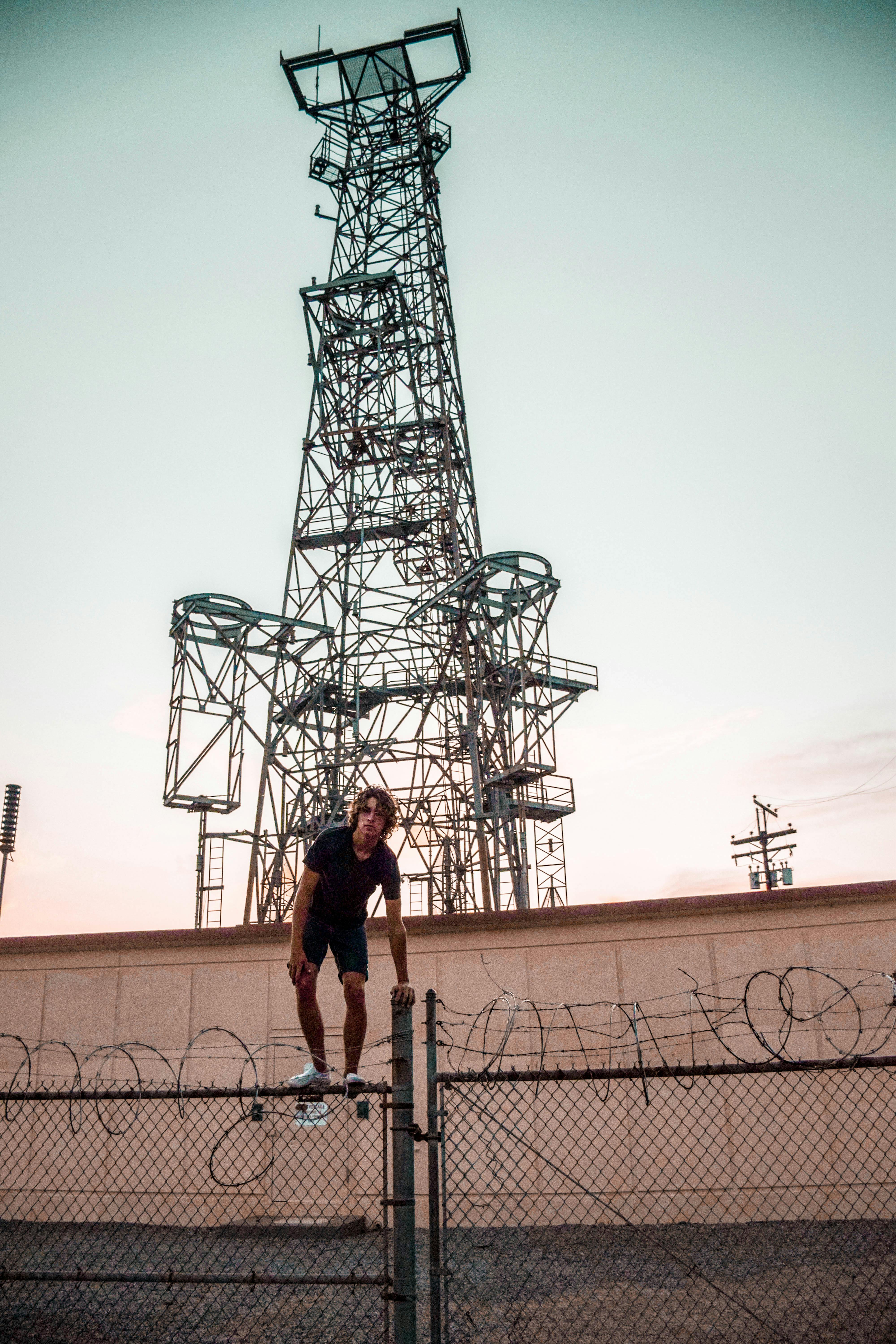 A Man Climbing Over A Barbed Wire Fence Near A Large Transmission Tower At Dusk.