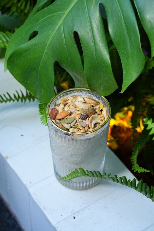 Free Yogurt glass topped with mixed nuts and seeds placed beside lush greenery featuring a monstera leaf. Stock Photo