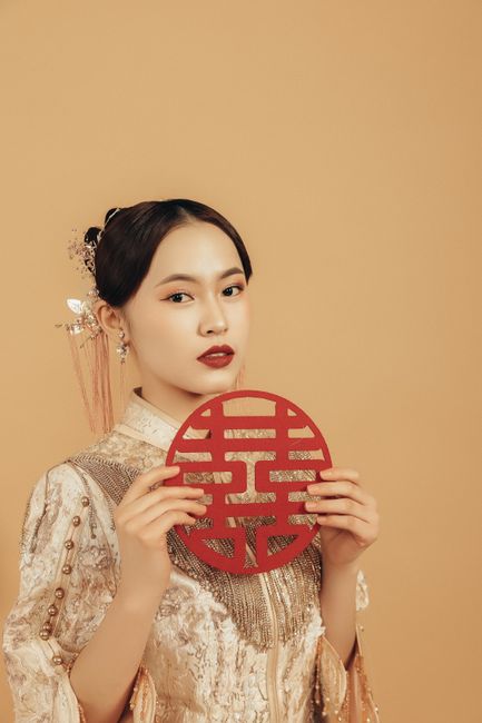 A young Asian woman elegantly dressed holds a red symbol of happiness against a neutral backdrop.