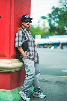 Street fashion portrait of a young woman leaning against a red wall wearing sunglasses and casual attire.