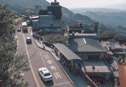 Aerial view of a bus stop with cars and people in a mountain village. Sunlit street and distant hills create a tranquil scene.