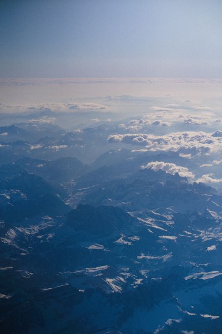A breathtaking aerial view showcasing snow-covered mountains under a clear blue sky with scattered clouds.