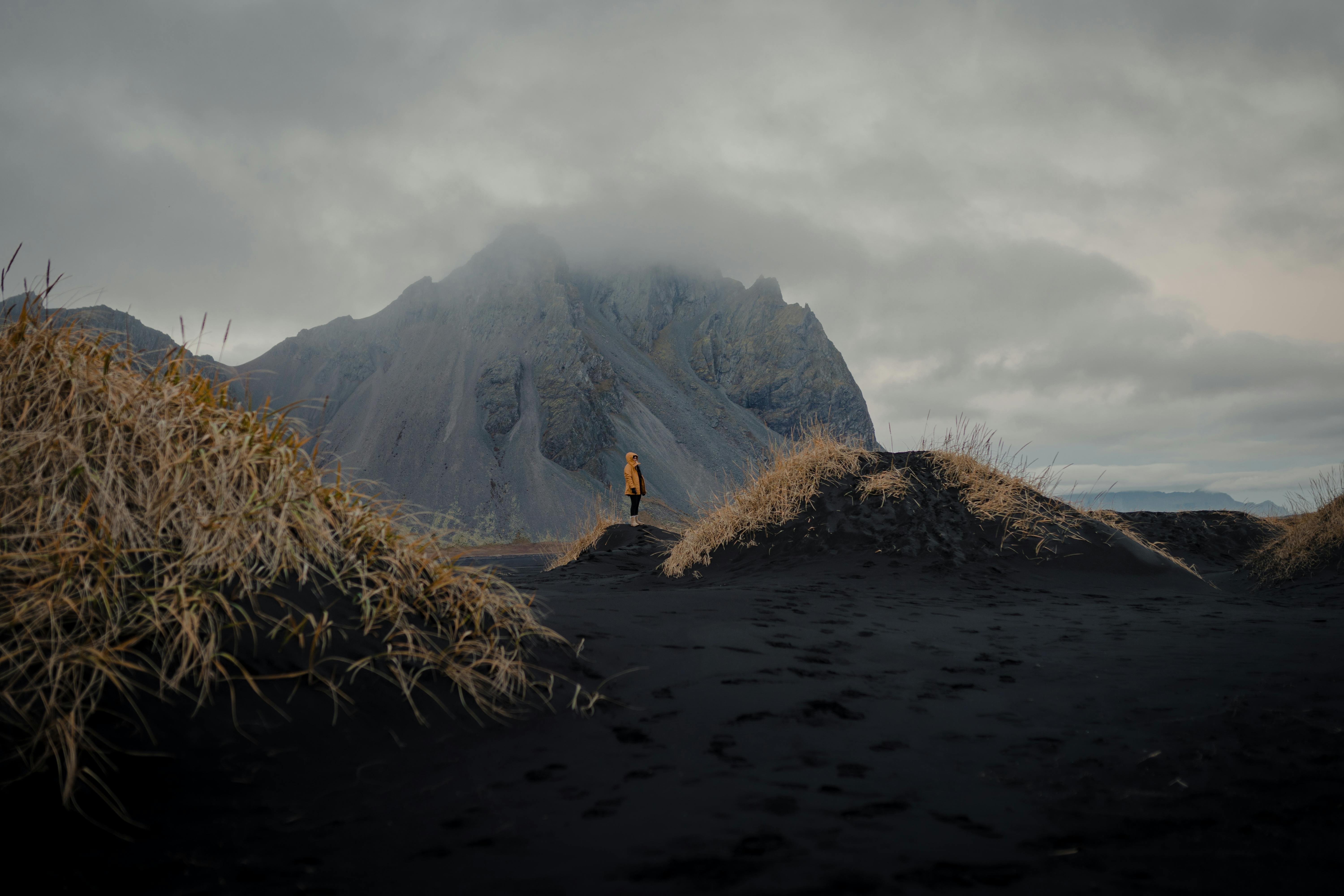 A solitary figure stands against a dramatic Icelandic mountain under a cloudy sky.