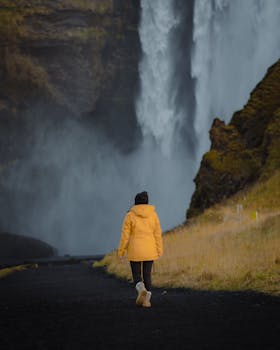 A person in a yellow raincoat walks towards a massive waterfall on a misty day.