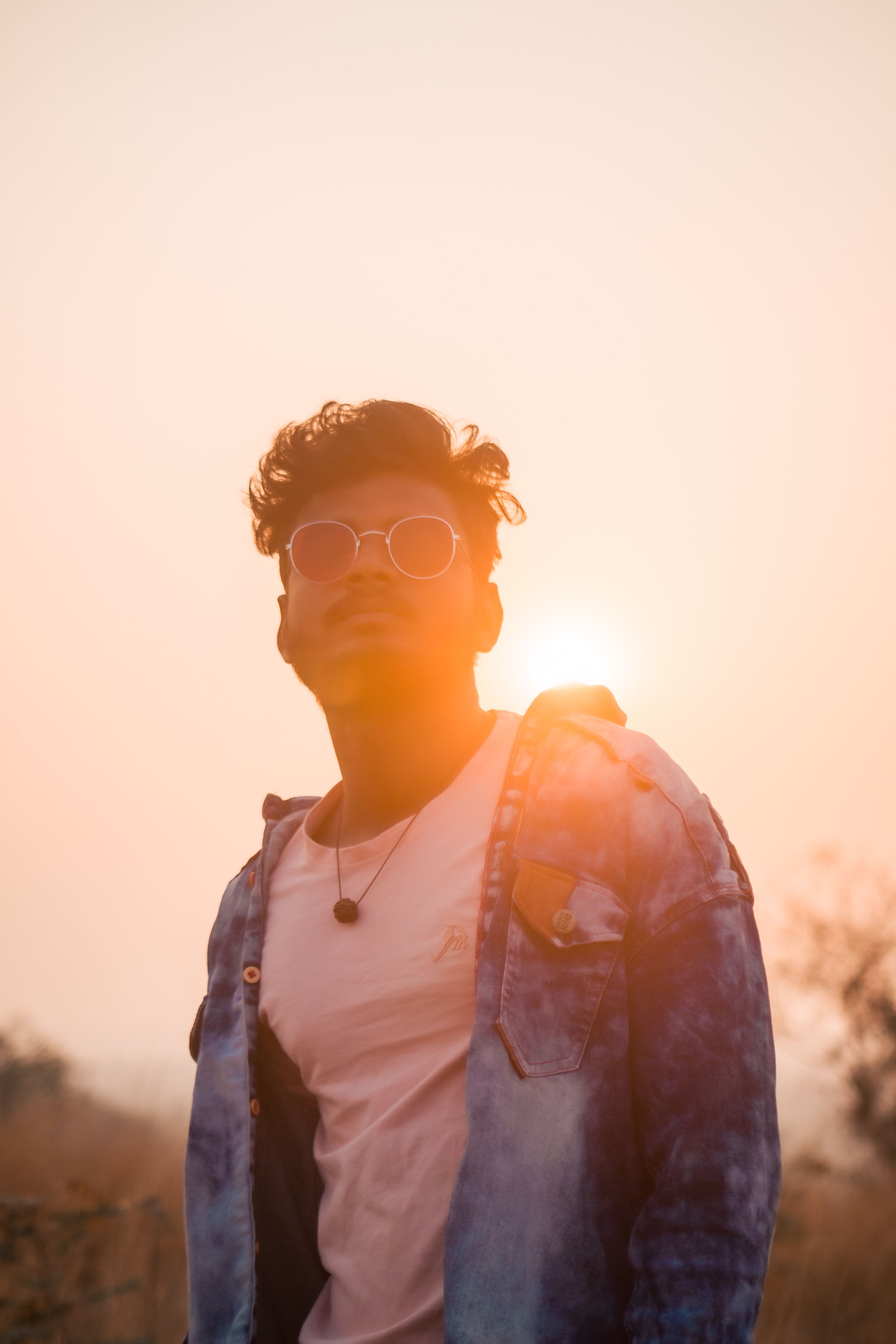 A fashionable man wearing sunglasses poses confidently at sunset, creating an artistic silhouette.