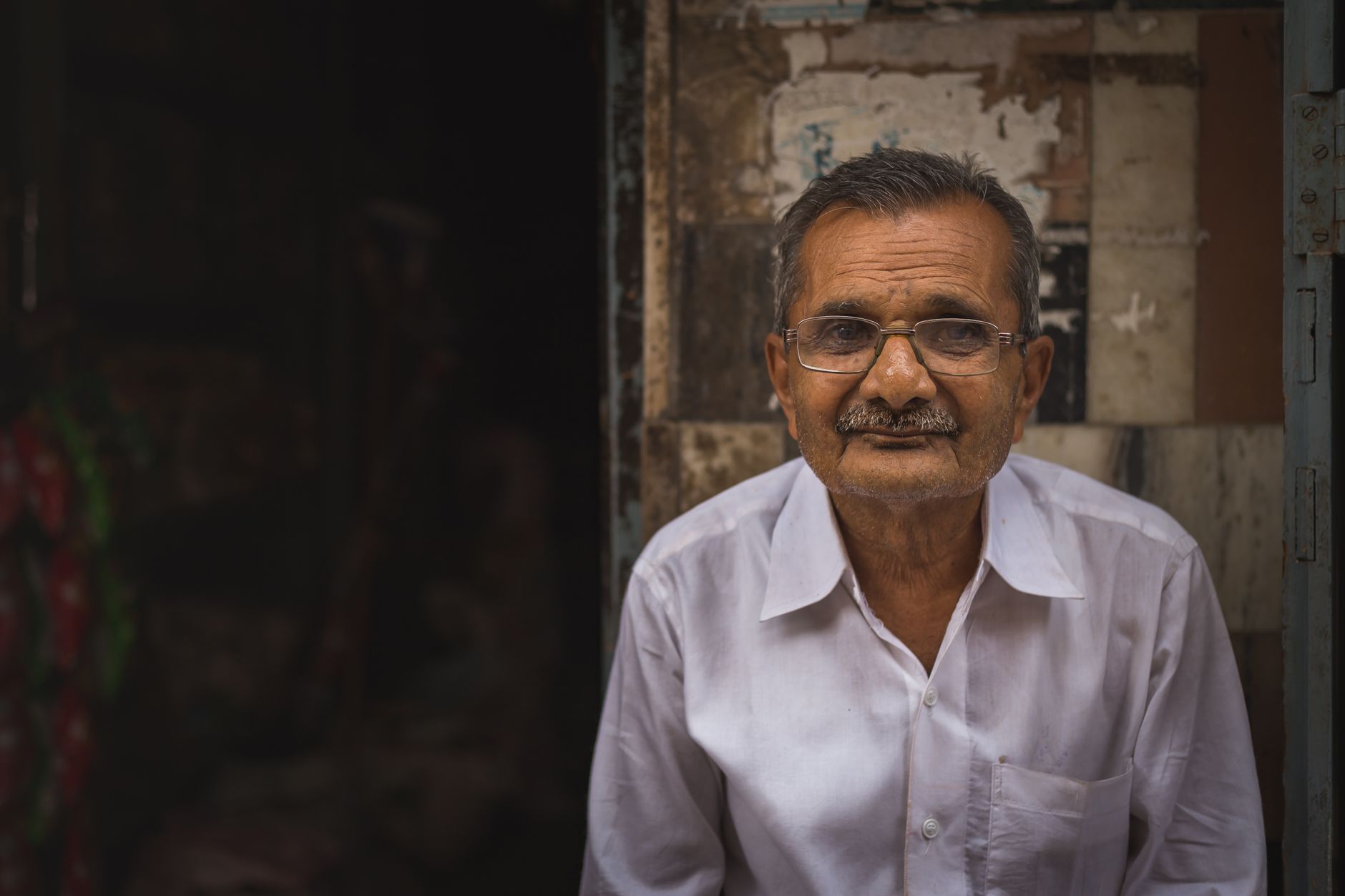 Elderly man with eyeglasses and a mustache in a white shirt, seated outdoors.