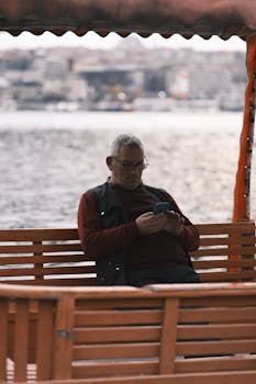Middle-aged man using cellphone on a bench by the water on a gloomy day.