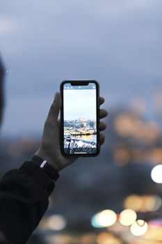 A hand holding a smartphone capturing a city skyline at dusk. Blurred lights in background.