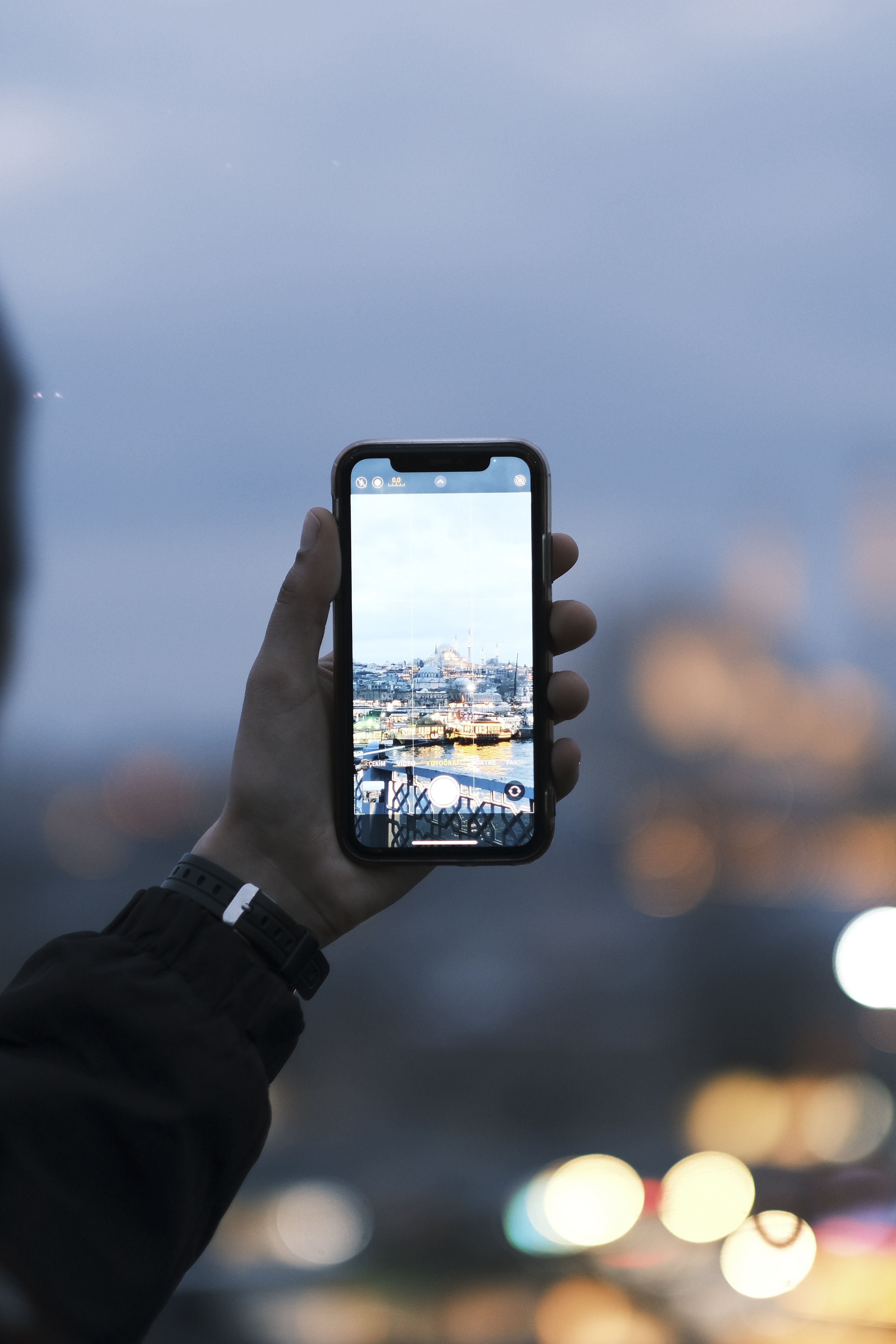 Free A hand holding a smartphone capturing a city skyline at dusk. Blurred lights in background. Stock Photo