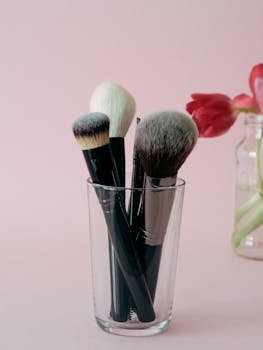 Stylish display of makeup brushes in a glass against a soft pink background with delicate flowers.