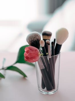 Close-up of makeup brushes and pink tulip in glass on a soft background.