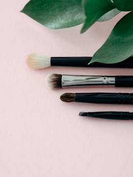 Aesthetic flat lay of makeup brushes with green leaves on a soft pink background.