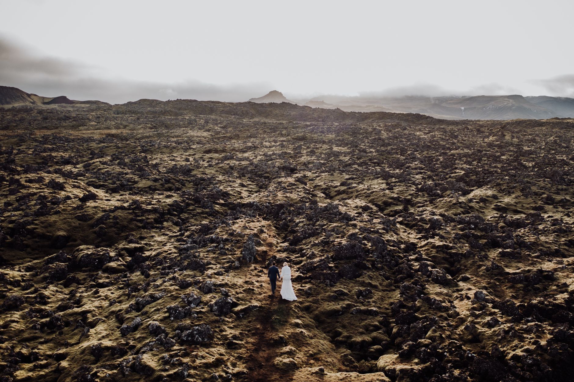 Couple in wedding attire walking across a vast, rocky Icelandic landscape at sunset.