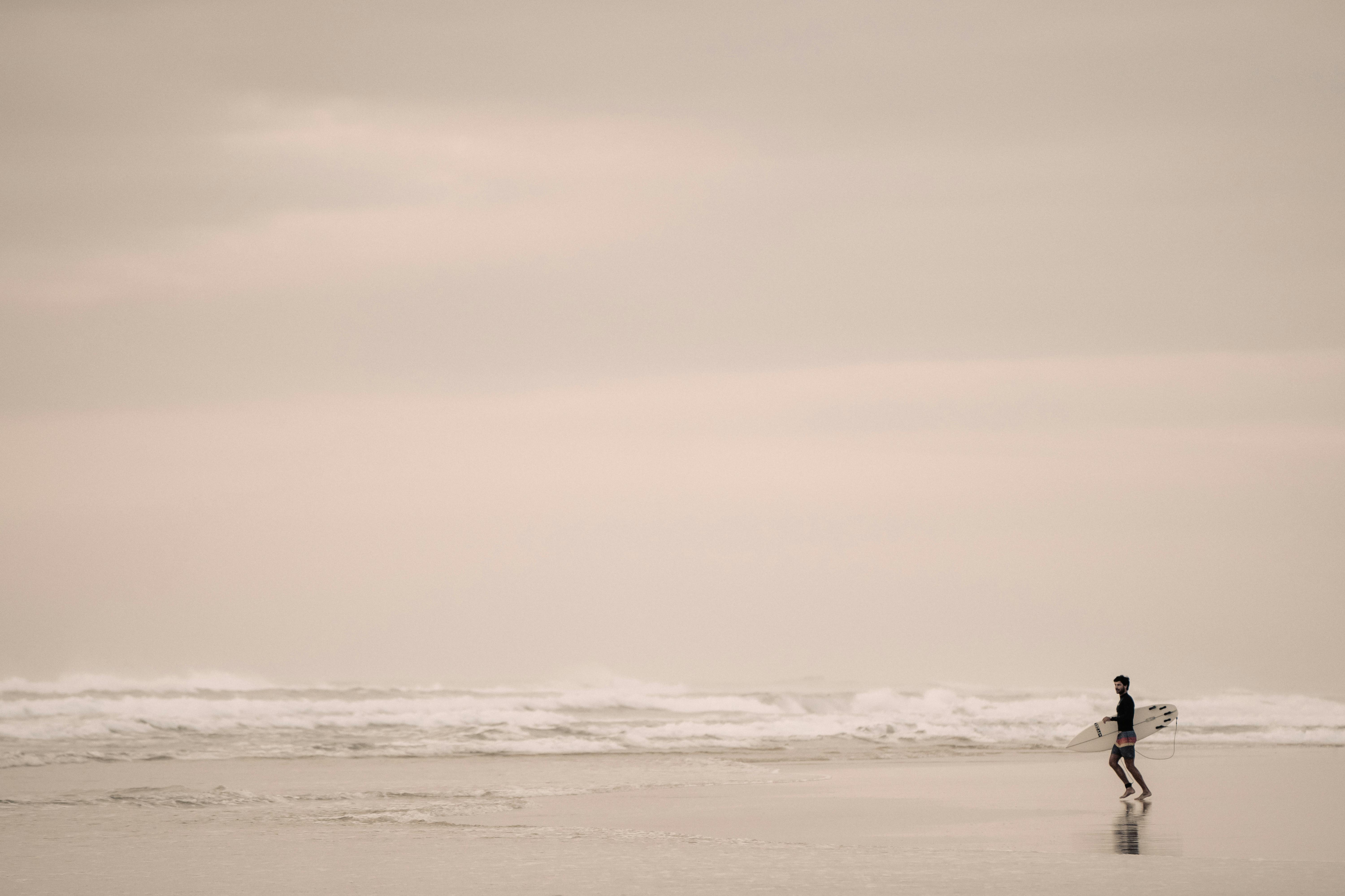 A surfer walks along Sunshine Beach at sunrise, ready to catch waves in the tranquil morning setting.
