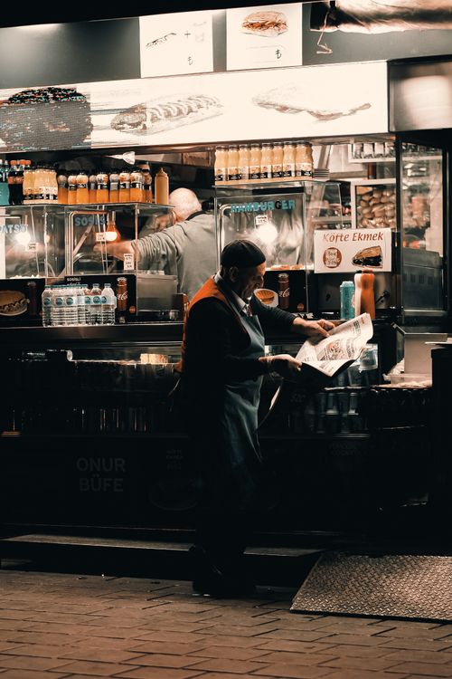Free An elderly man reads a newspaper at a vibrant street food stall in the evening. Stock Photo