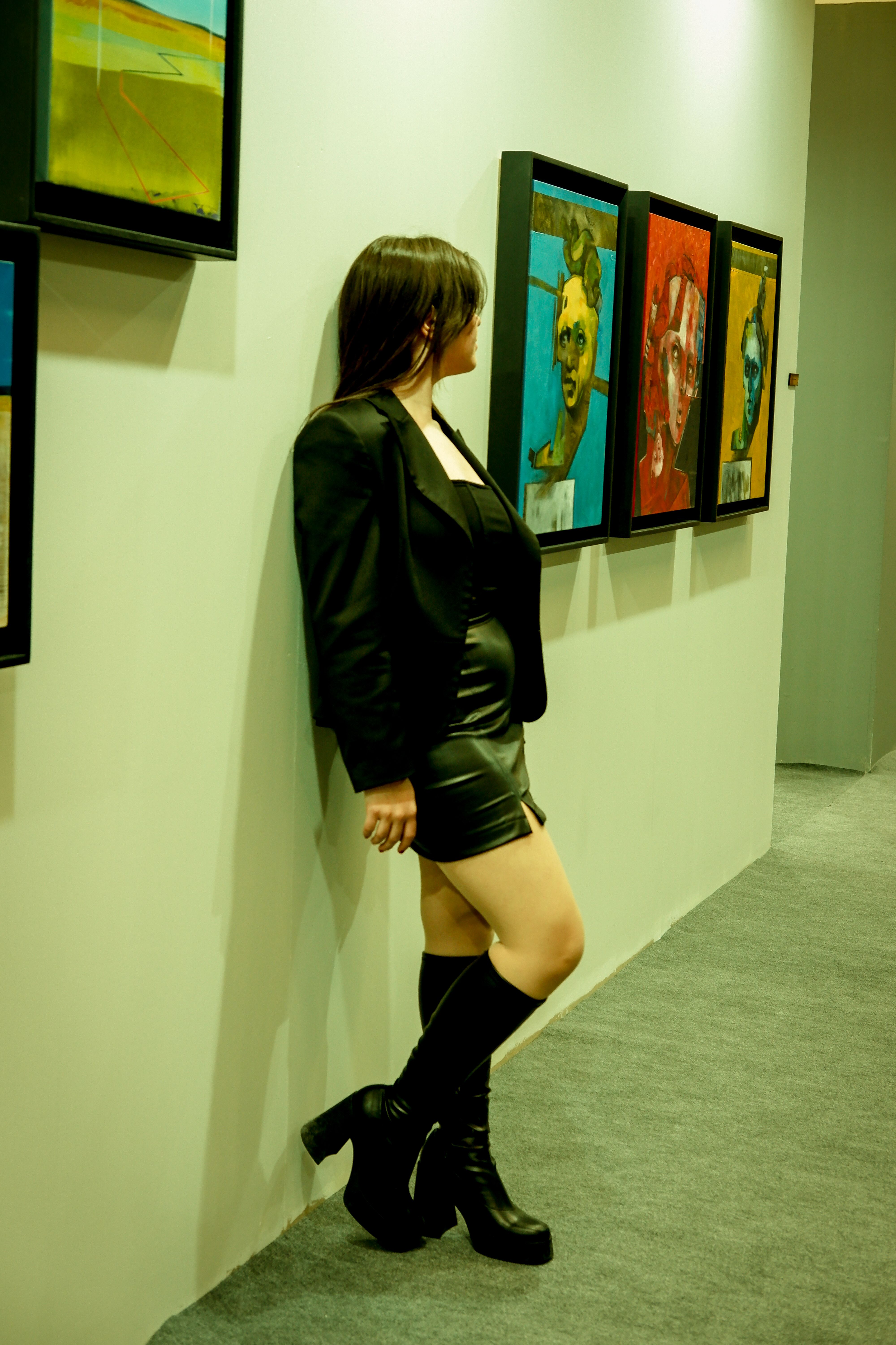 Stylish woman in a black jacket and platform heels observing paintings in an art gallery.