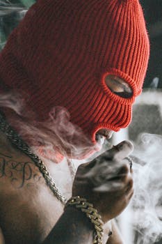 A person wearing a red balaclava smoking, with visible tattoos and gold chain, in an urban environment.