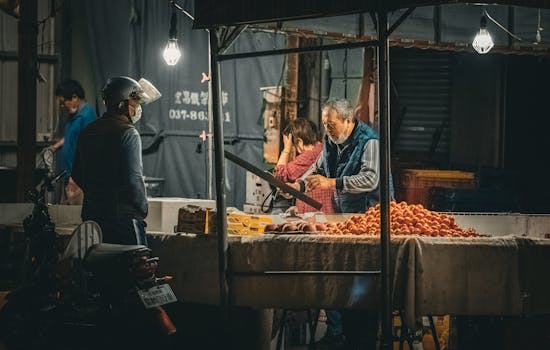 A night market vendor sells fresh produce under hanging lights, engaging with a customer.