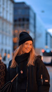 Woman in winter attire smiles warmly at dusk on a city street, showcasing modern winter fashion.