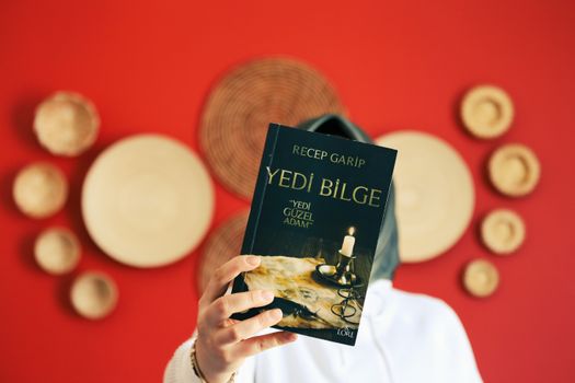 Person holding 'Yedi Bilge' book in front of a red wall with decorative plates. Close-up focus on book.