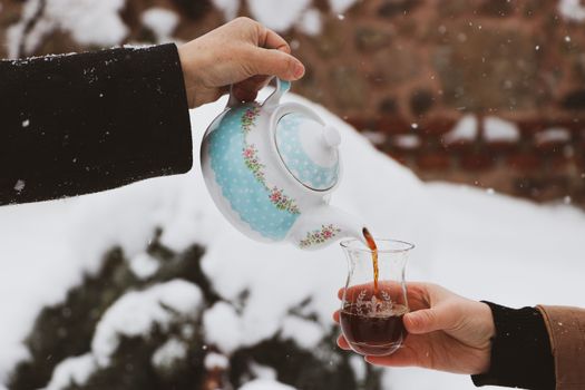 Close-up of hands pouring tea from a floral teapot into a glass in a snowy outdoor scene.