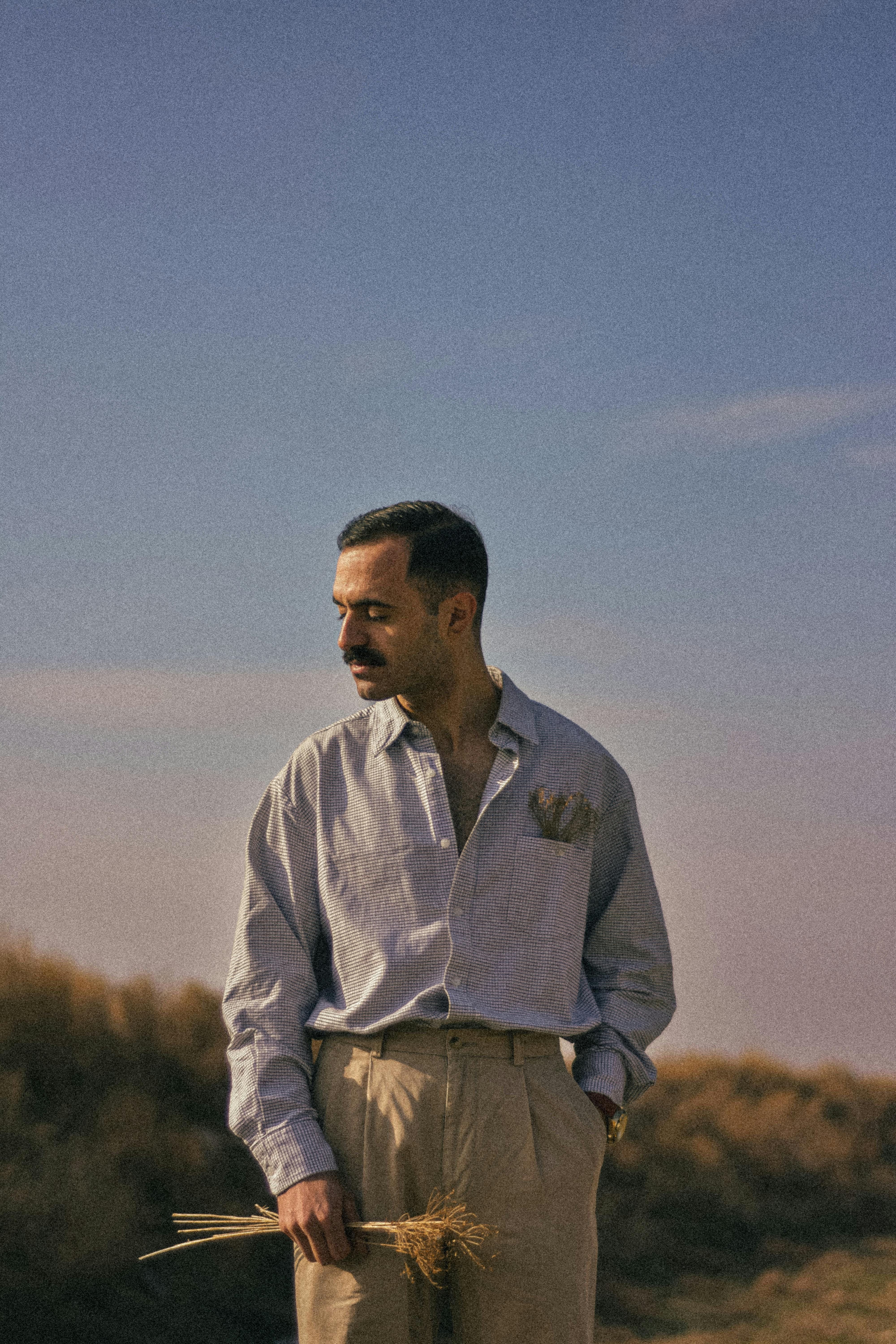 A man with a mustache holds wheat in a sunny field, wearing a shirt and trousers.