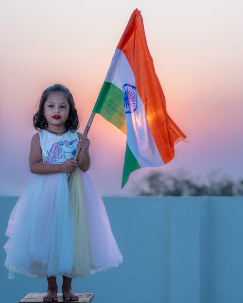 Free A young girl in a dress proudly holds the Indian flag at sunset, symbolizing patriotism. Stock Photo
