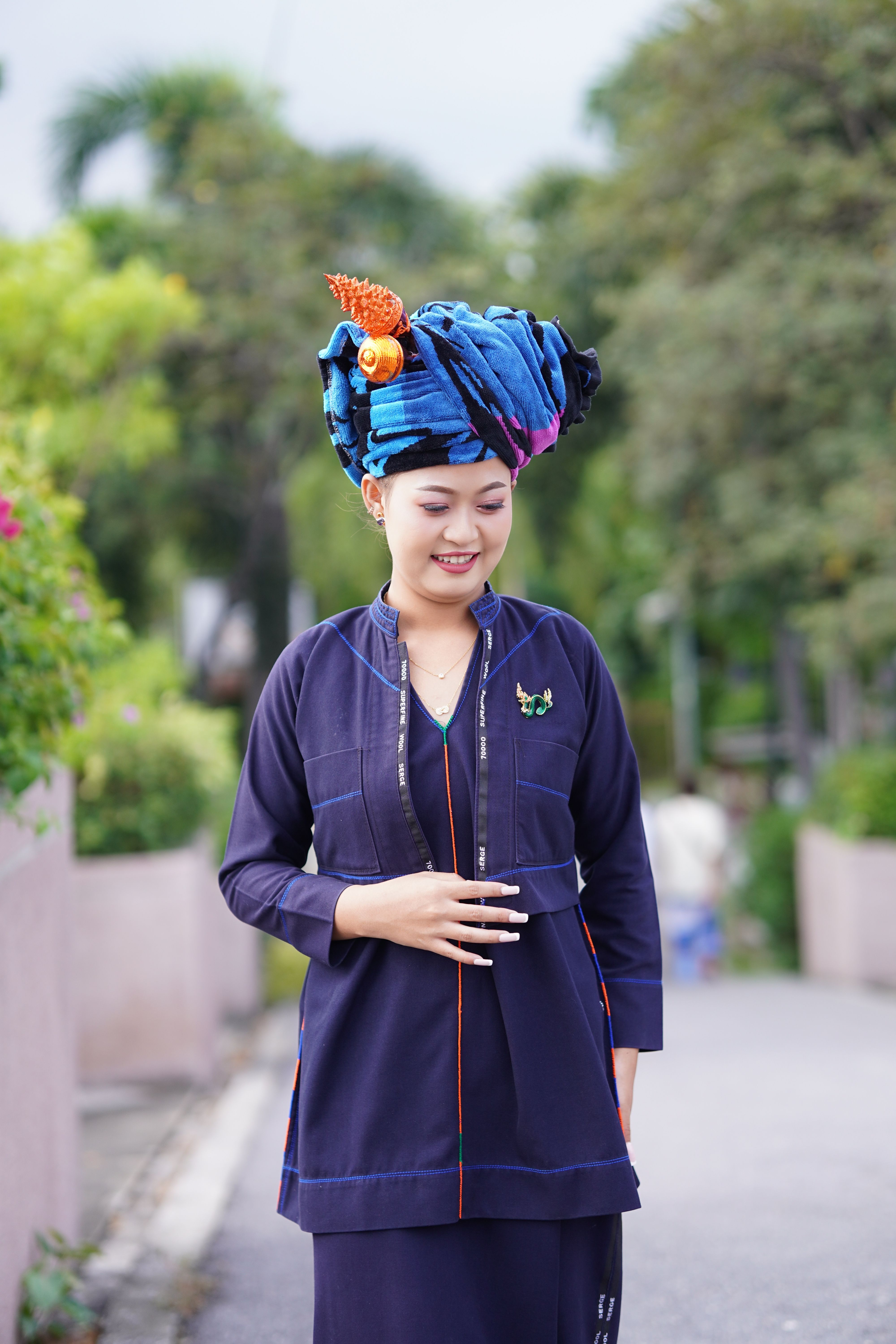 Smiling woman wearing a colorful turban and traditional dress, standing outdoors in a park setting.