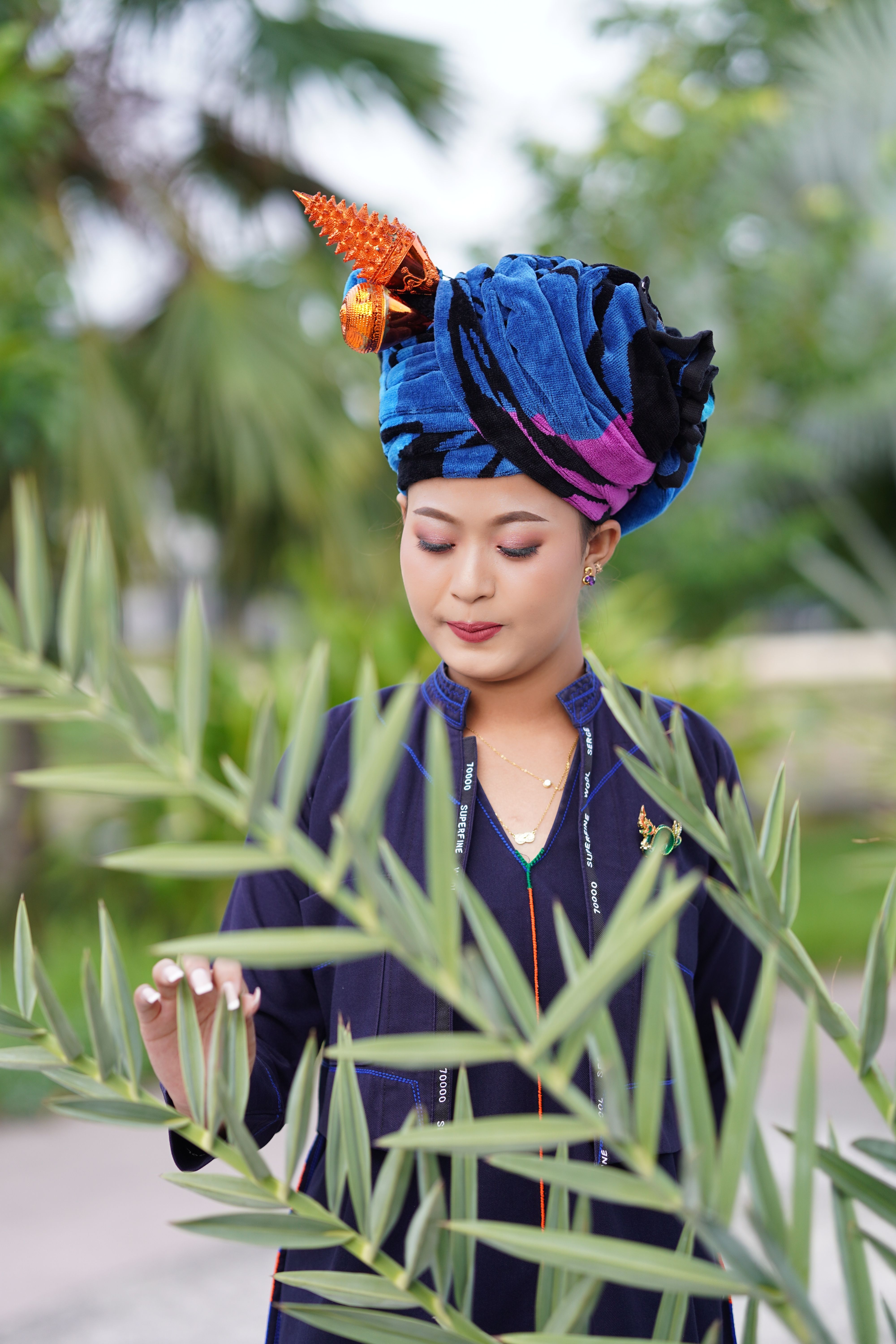 A young woman in a vibrant turban stands among green foliage outdoors.