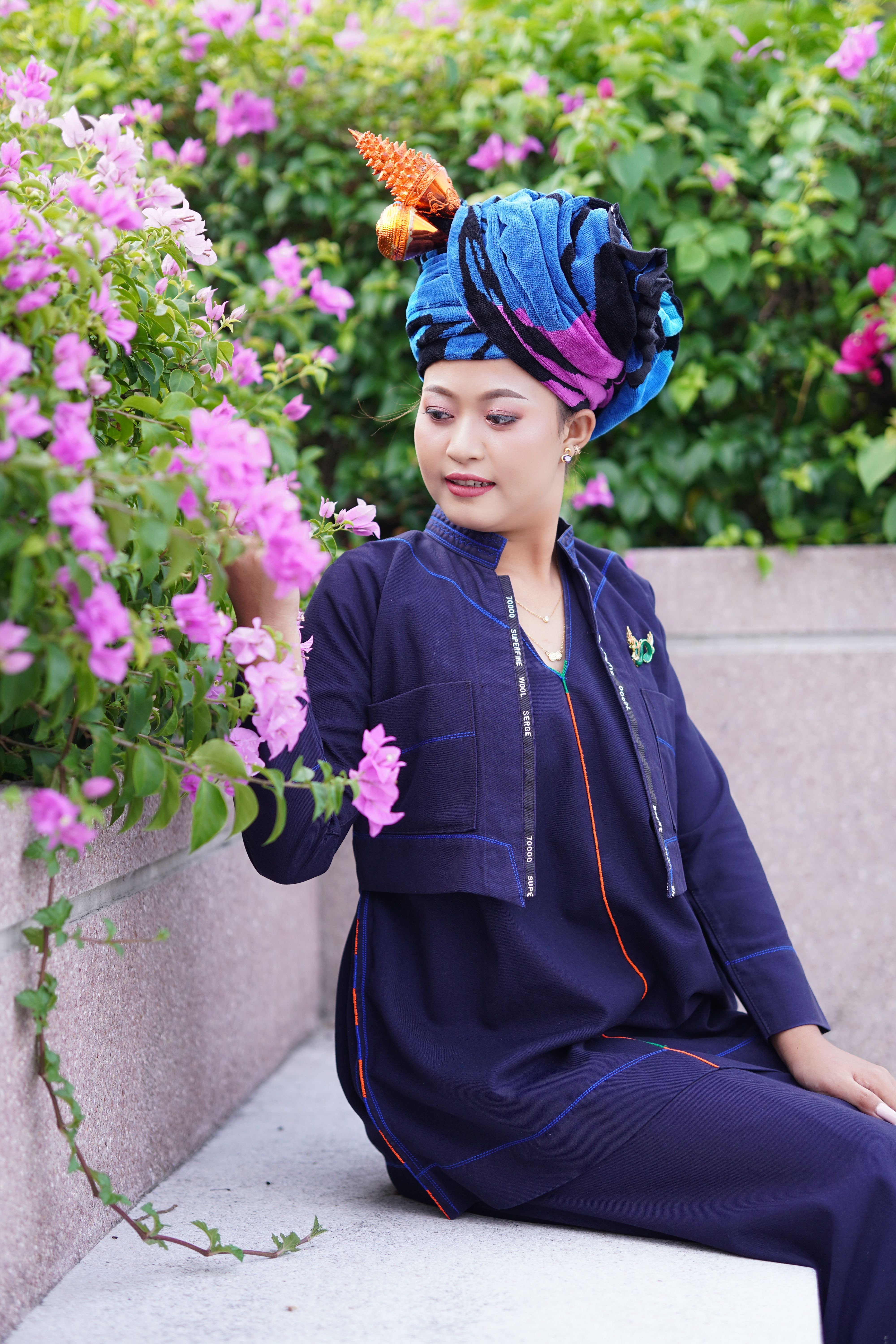Asian woman in vibrant turban sitting by flowers, embodying cultural grace.