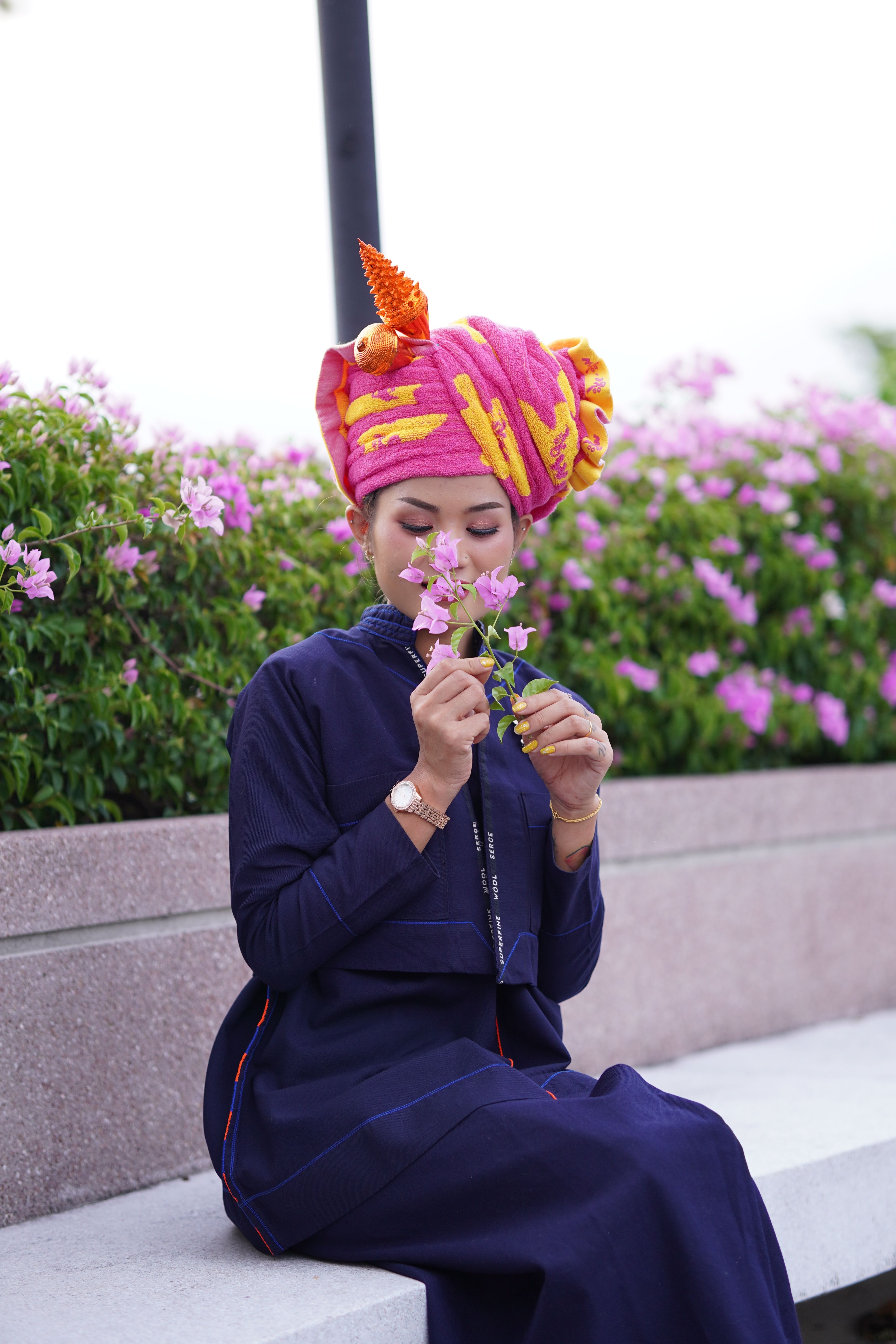 An Asian woman in traditional attire smells flowers while sitting on a bench outdoors.