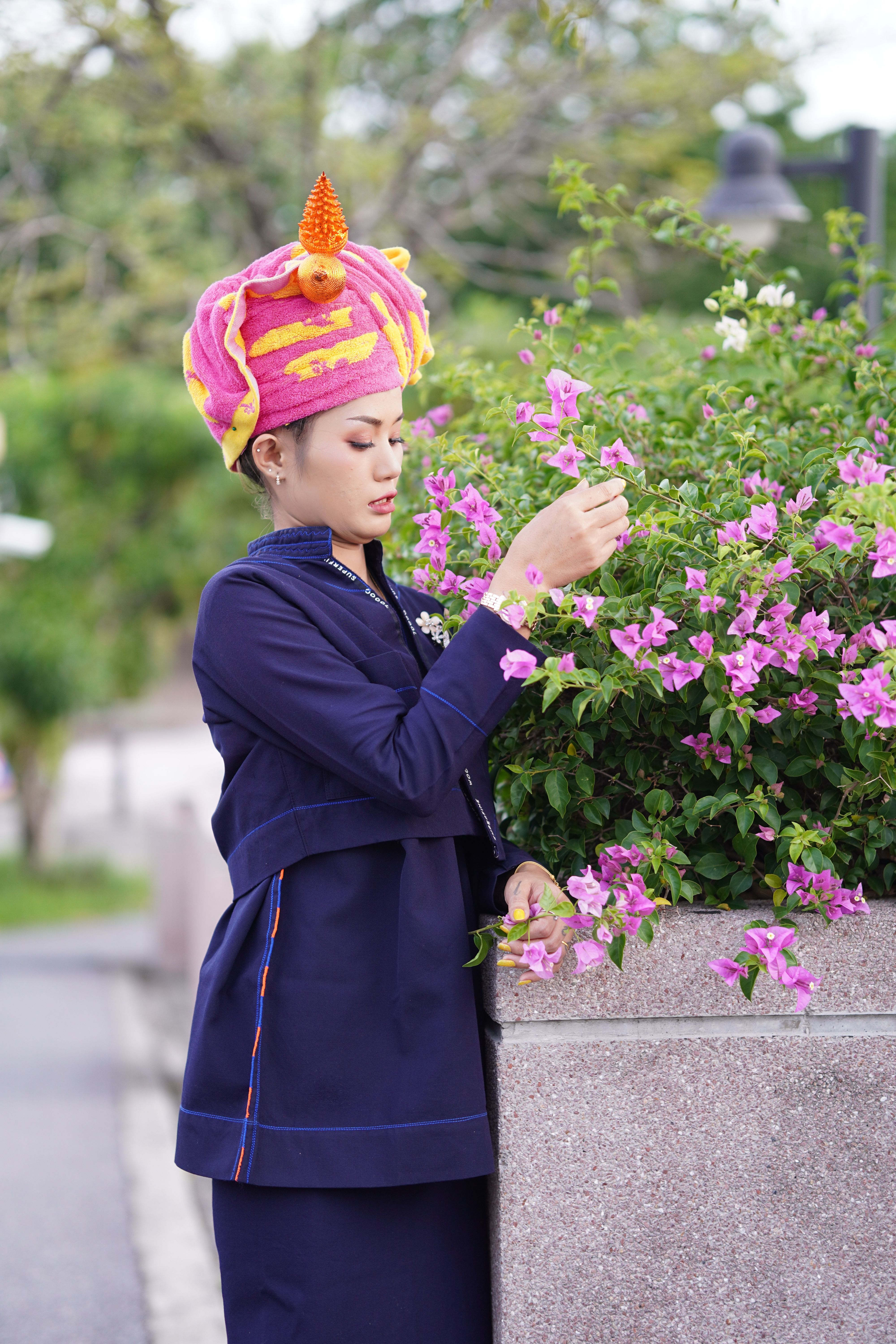 Portrait of a woman in traditional attire and colorful headdress, admiring flowers outdoors.