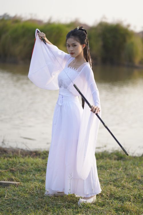 Free A woman in a white costume with a sword posing by a serene lakeside. Stock Photo
