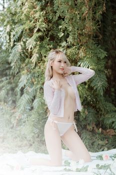 A young woman poses outdoors in lingerie, surrounded by lush green foliage.