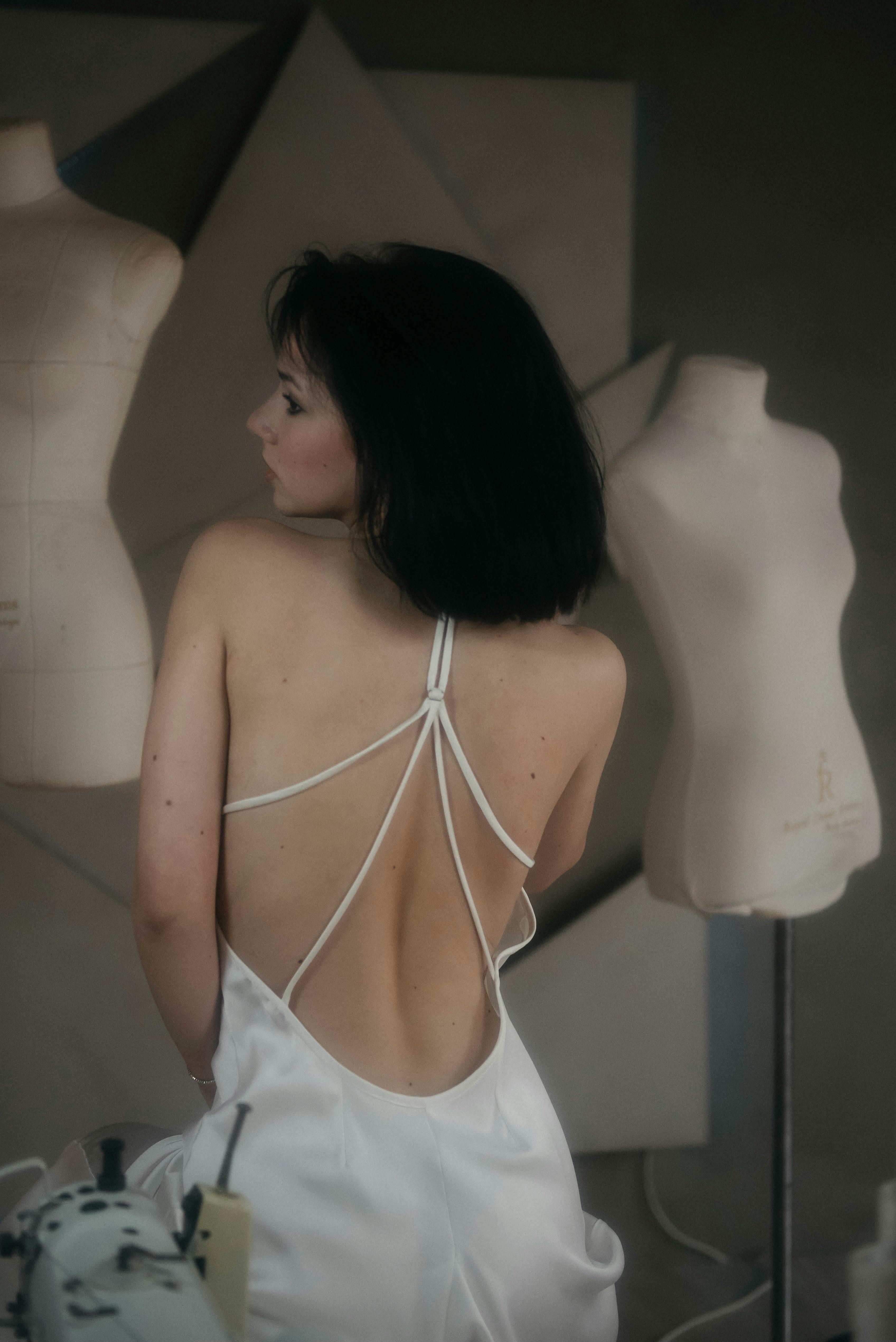 Model posing in a backless white dress surrounded by mannequins in a studio setting.