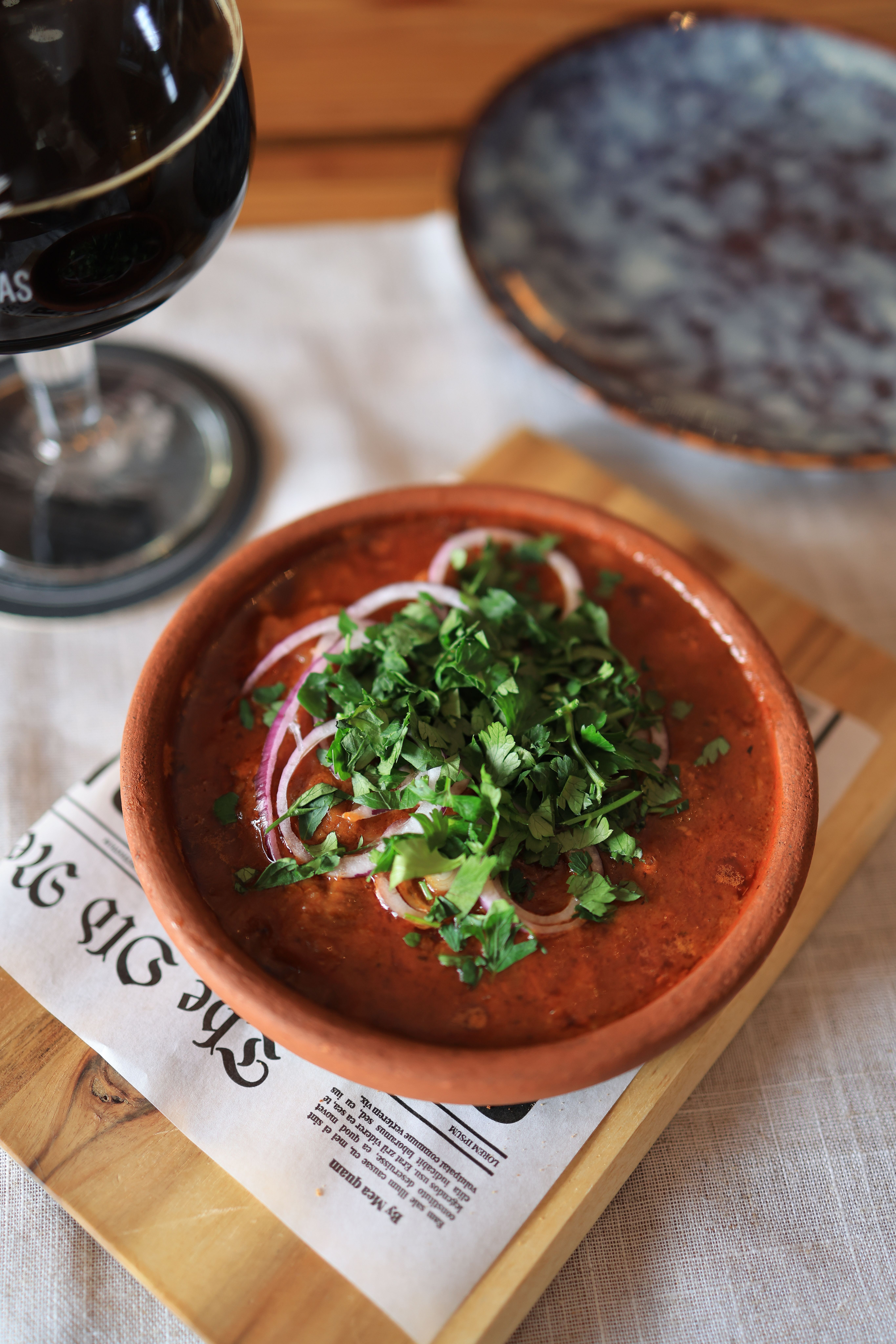 Close-up of a spicy soup garnished with fresh herbs served in a terracotta bowl with a glass of wine.